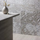 Large-format light gray marble tiles with natural marbled texture, shown on a wall and floor in a modern interior setting with a stone-look counter and a small vase of branches.