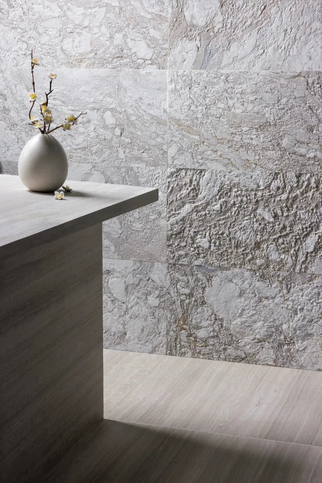 Large-format light gray marble tiles with natural marbled texture, shown on a wall and floor in a modern interior setting with a stone-look counter and a small vase of branches.