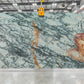 Large quartzite slab panel with a pale blue-green base and prominent dark gray veining plus warm orange-gold veining, photographed hanging in a warehouse or showroom.