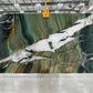 A large rectangular quartzite slab of polished green marble with dramatic veining, suspended by a yellow crane hook in a warehouse setting.