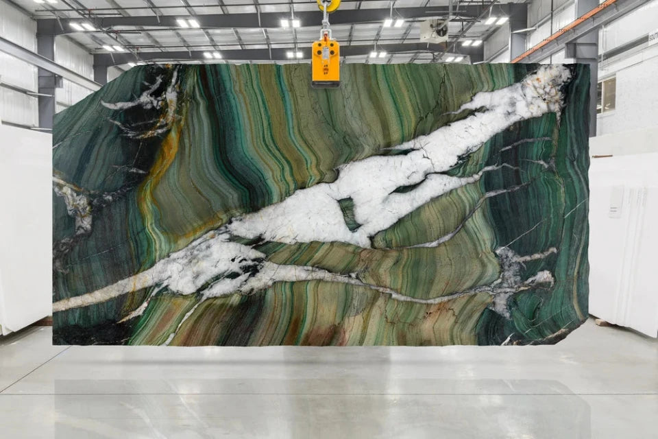 A large rectangular quartzite slab of polished green marble with dramatic veining, suspended by a yellow crane hook in a warehouse setting.