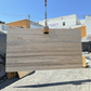 A large rectangular stone slab with natural beige, brown and taupe veining is suspended by a crane chain outdoors in los angeles slab yard on a concrete surface, with buildings and a blue sky in the background.