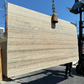 A large rectangular stone slab with natural beige, brown and taupe veining is suspended by a crane chain outdoors in los angeles slab yard on a concrete surface, with buildings and a blue sky in the background.