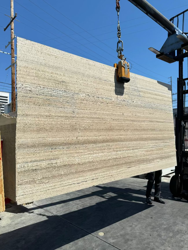 A large rectangular stone slab with natural beige, brown and taupe veining is suspended by a crane chain outdoors in los angeles slab yard on a concrete surface, with buildings and a blue sky in the background.