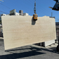 A large rectangular slab of light beige travertine stone slab is suspended by a crane hook in an outdoor los angeles slab yard under a blue sky.