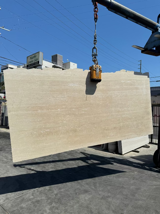 A large rectangular slab of light beige travertine stone slab is suspended by a crane hook in an outdoor los angeles slab yard under a blue sky.