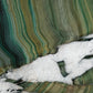 A close-up of a polished quartzite stone with wavy bands of green, teal, olive, beige, and white, including a prominent white fractured area running diagonally across the lower middle.